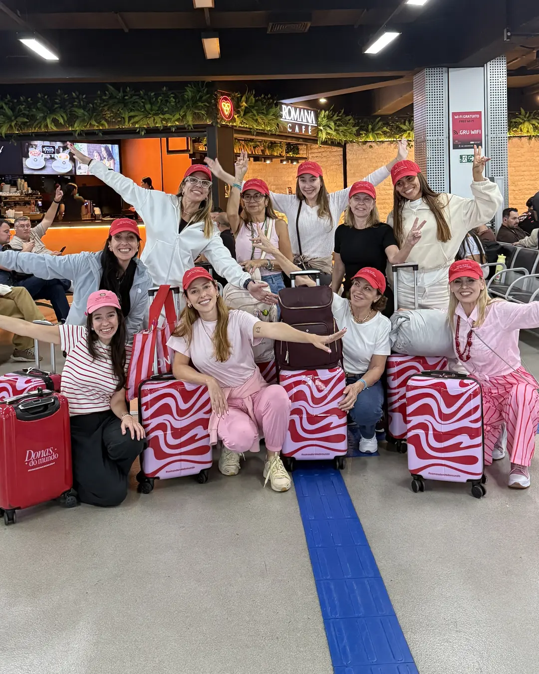 Grupo de Donas de boné vermelho comemorando no aeroporto, com as malas listradas de vermelho e rosa da Donas do Mundo