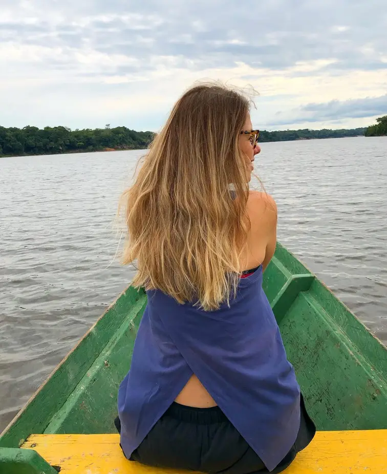 Mulher de cabelos longos, de costas, sentada na ponta de uma canoa verde olhando o rio