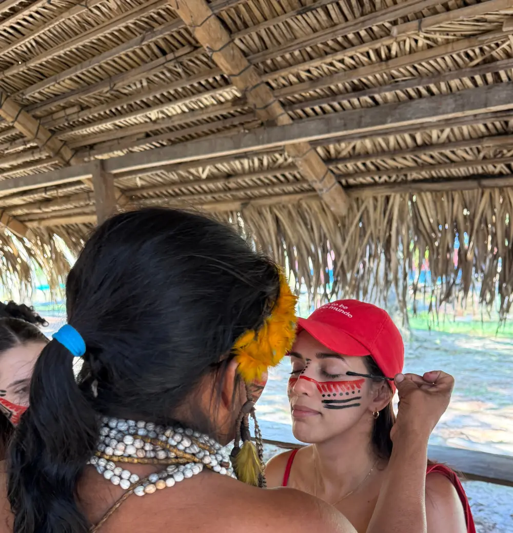 Mulher indígena fazendo pintura no rosto de uma Dona de boné vermelho