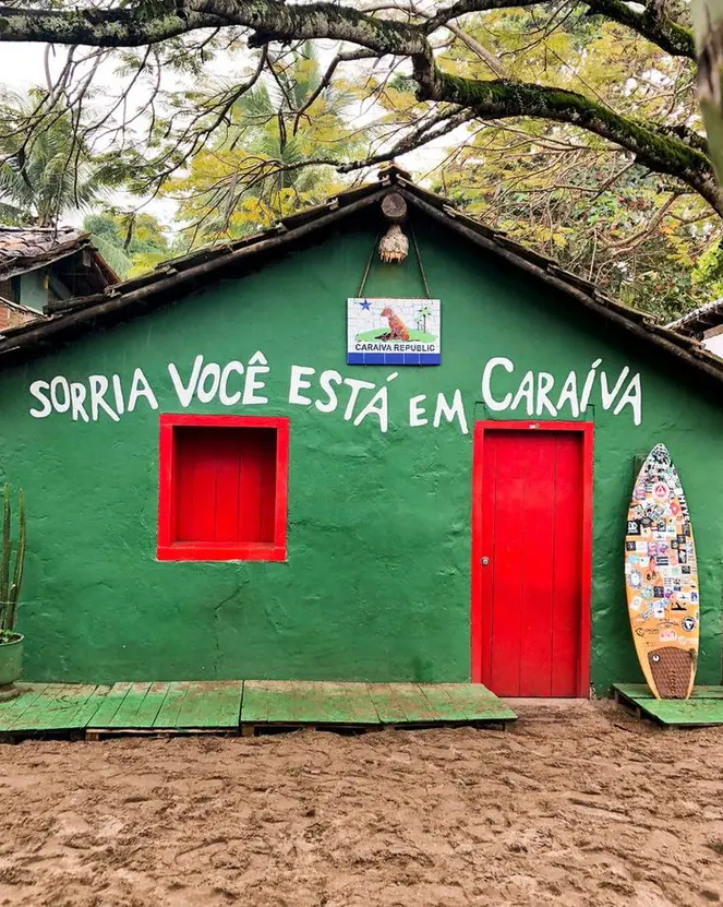 Casinha verde com porta e janela vermelhas e a frase “Sorria, você está em Caraíva” pintada na fachada
