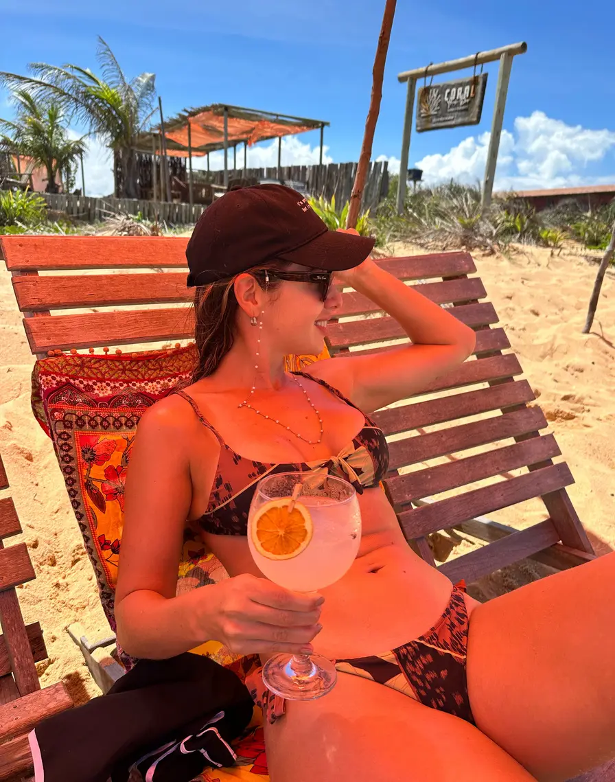 Dona de boné e biquíni brindando com um drink em uma espreguiçadeira na praia