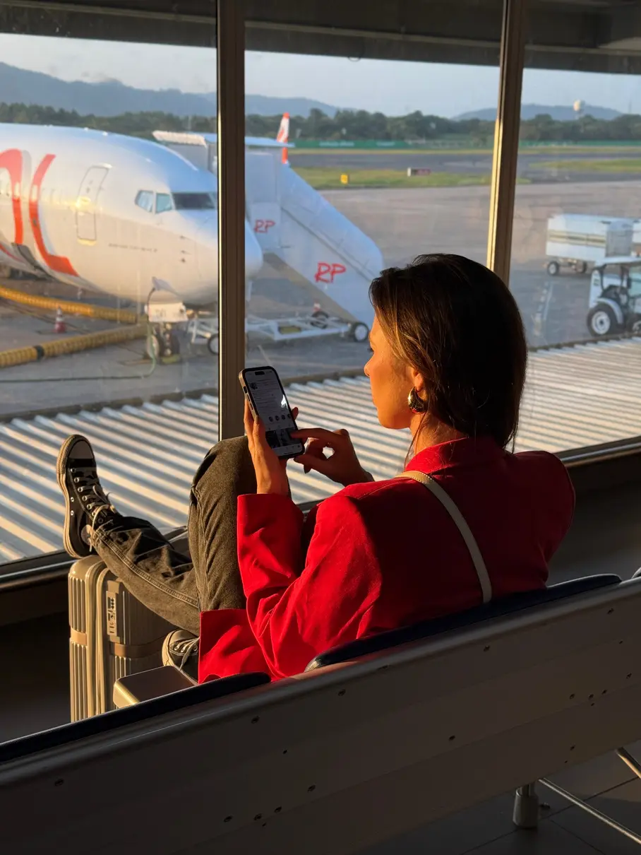 Mulher de casaco vermelho no saguão do aeroporto, olhando o celular diante da janela com aviões