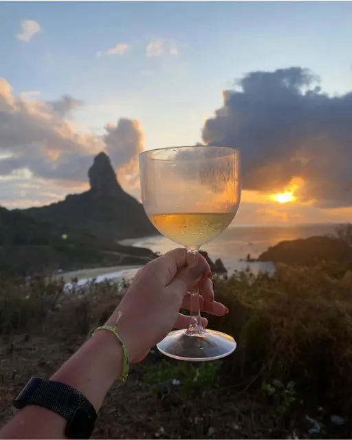 Mão segurando uma taça de vinho branco diante do pôr do sol em Noronha