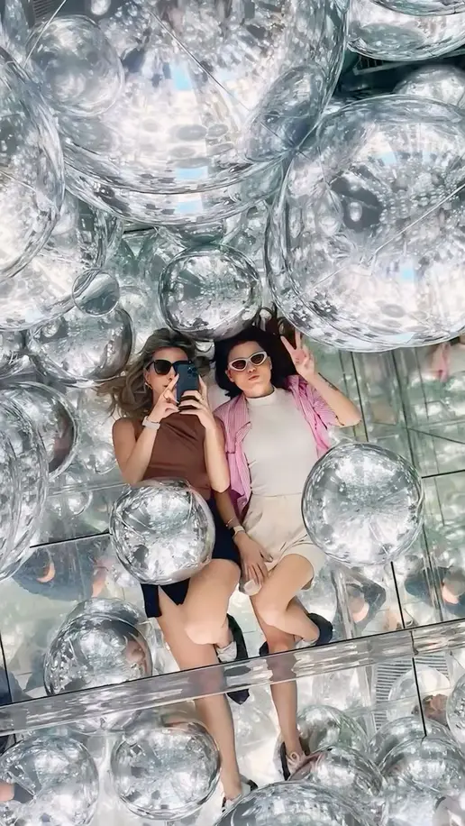 Duas amigas deitadas entre esferas espelhadas em uma instalação artística