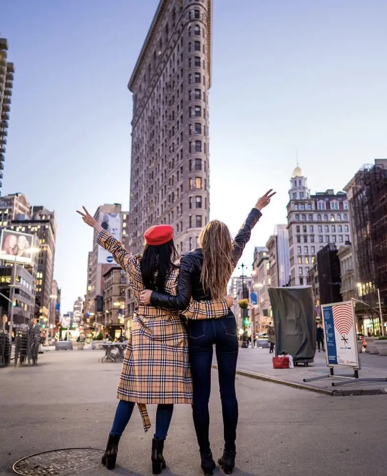 Duas amigas de braços erguidos em uma avenida de Nova York, com o Flatiron Building ao fundo
