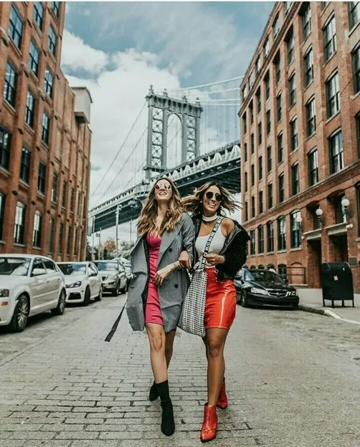 Duas amigas caminhando por uma rua de paralelepípedos em DUMBO, com a Ponte de Manhattan ao fundo