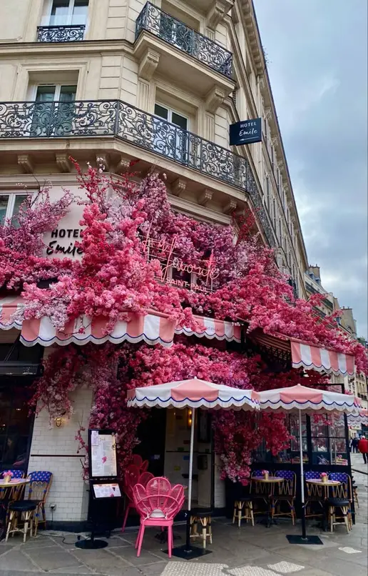 Fachada de café parisiense coberta de flores cor-de-rosa, com guarda-sóis listrados